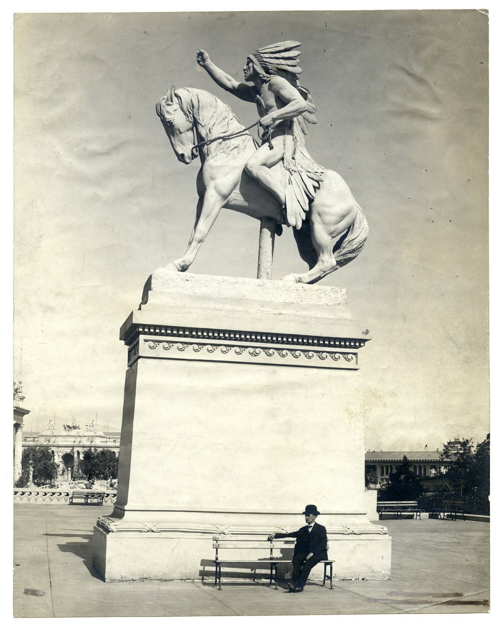 Verticaal, sepiafoto van een standbeeld van een Amerikaanse Indiaan zittend op een paard op een groot voetstuk. Er zit een man in een donker pak en hoed op een bankje aan de voet van het voetstuk door Unknown