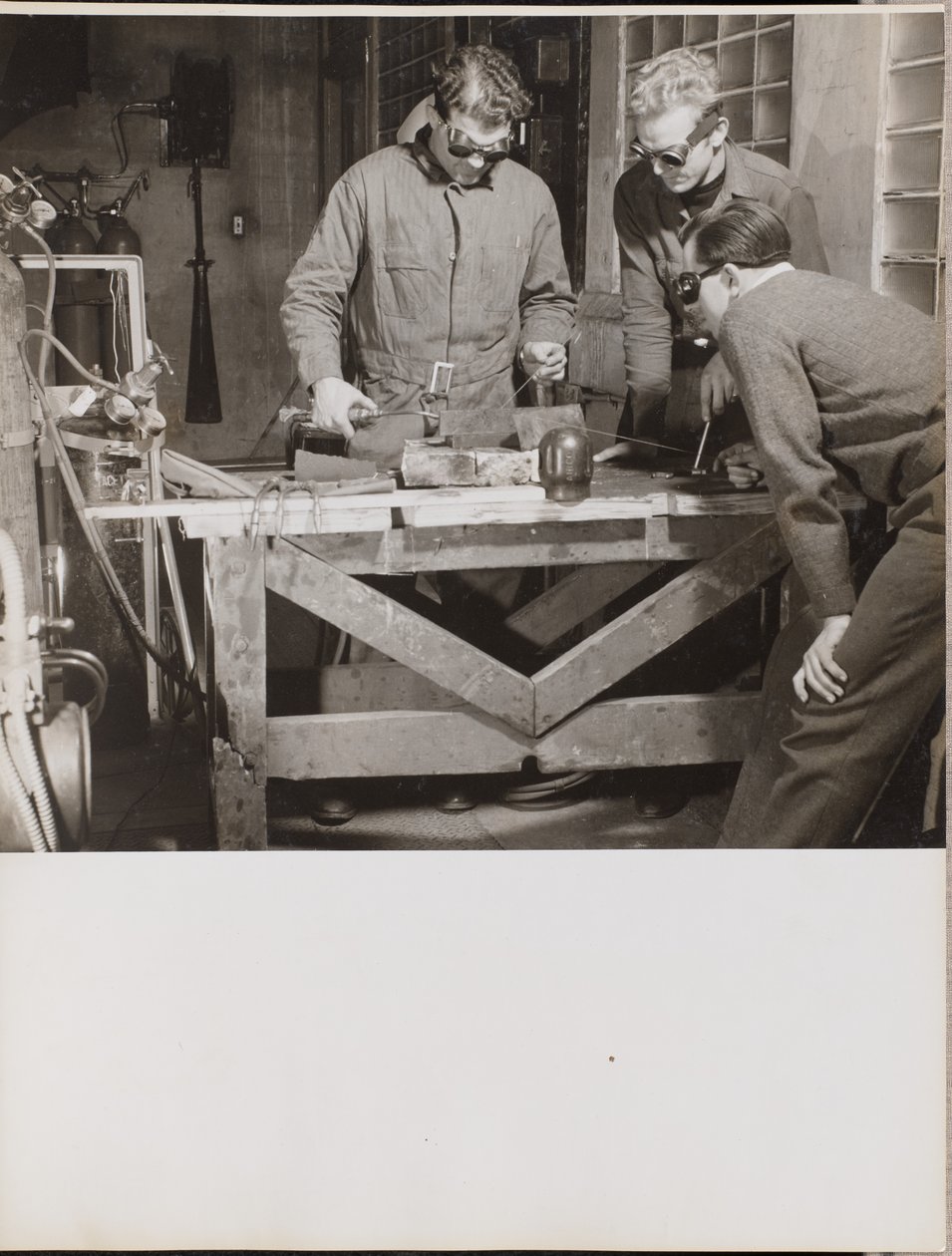 [Drie mannen met veiligheidsbrillen lassen een stuk metaal op een tafel]. door Arnold Eagle
