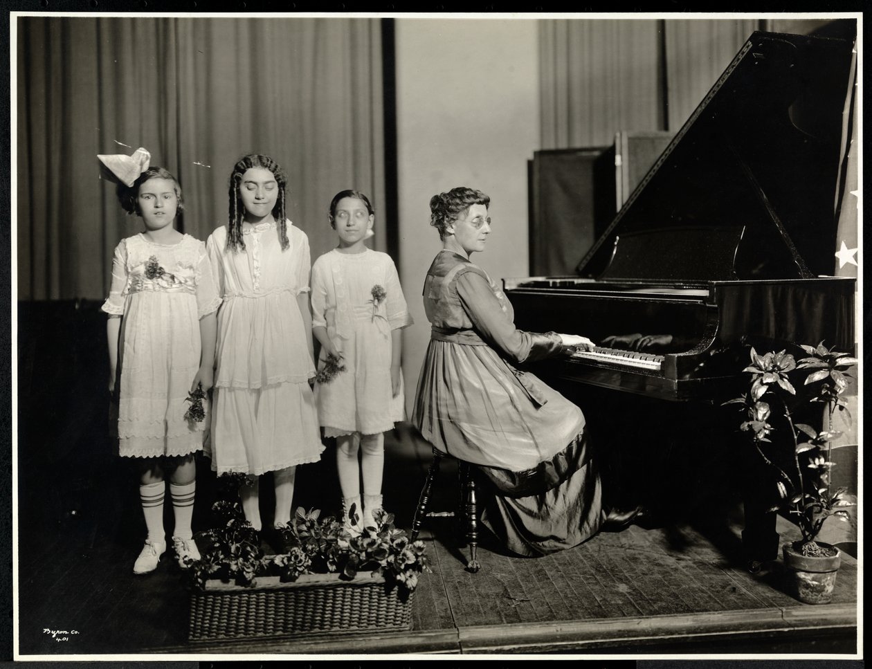 Vrouw speelt piano terwijl drie meisjes zingen bij de New York Association for the Blind, 111 East 59th Street, New York, c.1917 door Byron Company