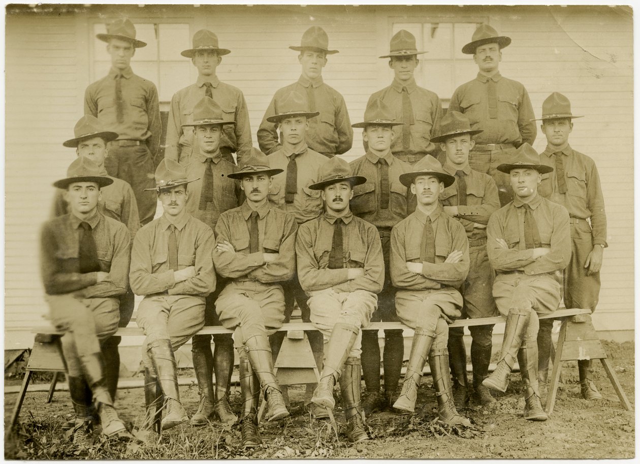 Horizontale sepia foto van een groep van zeventien uniformen soldaten die poseren voor een groepsfoto. De soldaten zijn verdeeld in drie rijen; een rij zittend vooraan, een rij staand in het midden en een rij staand op een platform achteraan door Carl Michel
