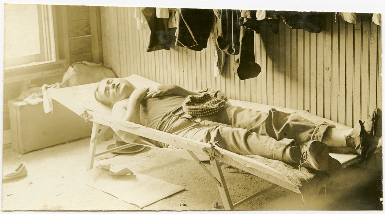 Horizontale, sepia foto van een man die slaapt op een veldbed in een houten gebouw. Diverse kledingstukken hangen aan de muur en koffers staan op de grond in de hoek van de kamer door Carl Michel