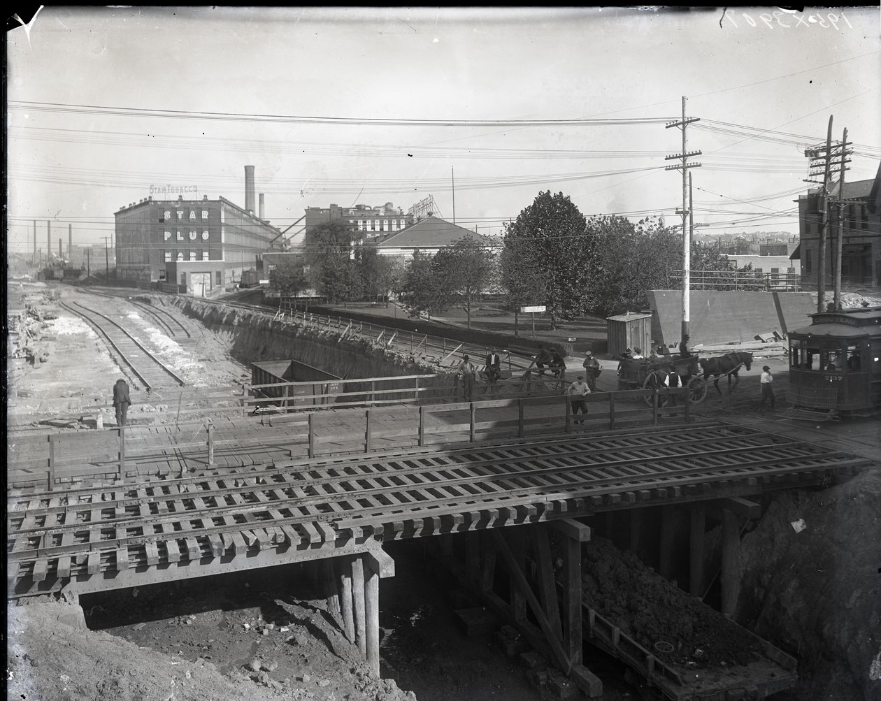 Horizontale zwart-witfoto van een houten brug en tramsporen over gedeeltelijk aangelegde spoorlijnen. Een tram gemarkeerd Chouteau 2137 begint de brug over te steken aan de rechterkant. In de verte fabrieksgebouwen met de tekst Star door Charles Clement Holt