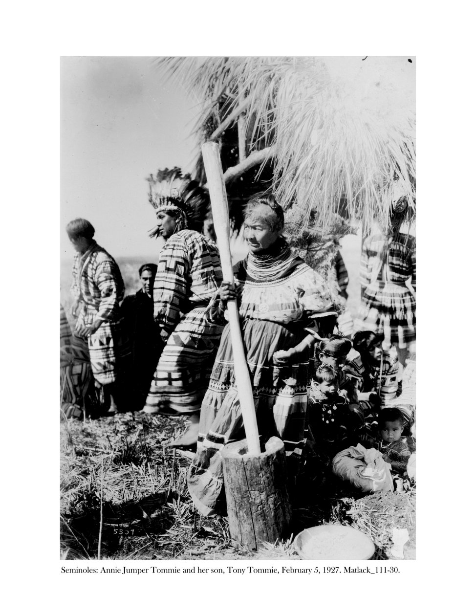 Annie Jumper Tommie gebruikt een vijzel en stamper om maïsmeel te maken, en haar zoon, Tony Tommie, draagt een adelaarsveren tooi, 5 februari, 1927 door Claude C. Matlack