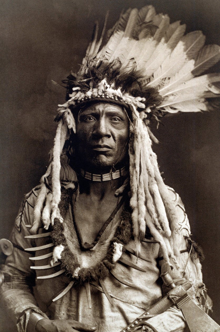Weasel Tail (Apohsuyis). Portret van Indiaanse hoofdman van de Piegan stam met de beroemde adelaarsveer en wezels tooi, een suède tuniek, twee kettingen, een van bot en de andere van grizzly klauwen, en een tomahawk pijp door Edward Sheriff Curtis