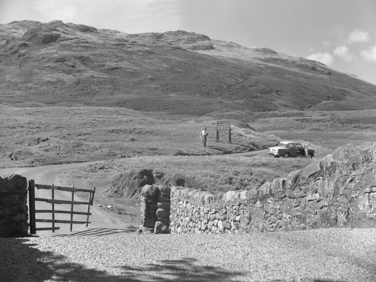 Zicht op de weg die over Hard Knott Pass gaat, twee mensen die bij een verkeersbord staan en een geparkeerde auto in de grasberm door Joseph Hardman