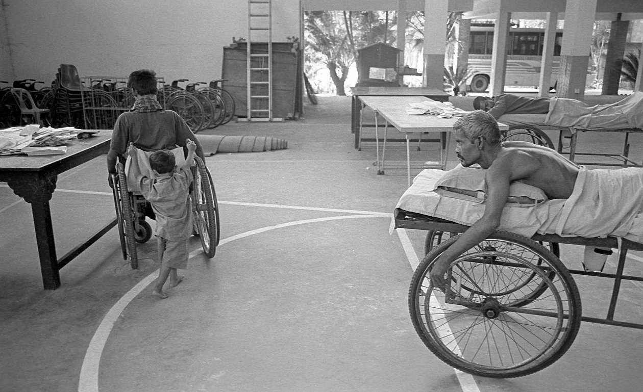 Een lichamelijk gehandicapte man in een rolstoel wordt gedragen door zijn zoon; een andere man met hetzelfde lot kijkt toe in het Centrum voor Revalidatie van Verlamden, CRP in Savar, Dhaka, Bangladesh. 2005. door Unbekannt Unbekannt