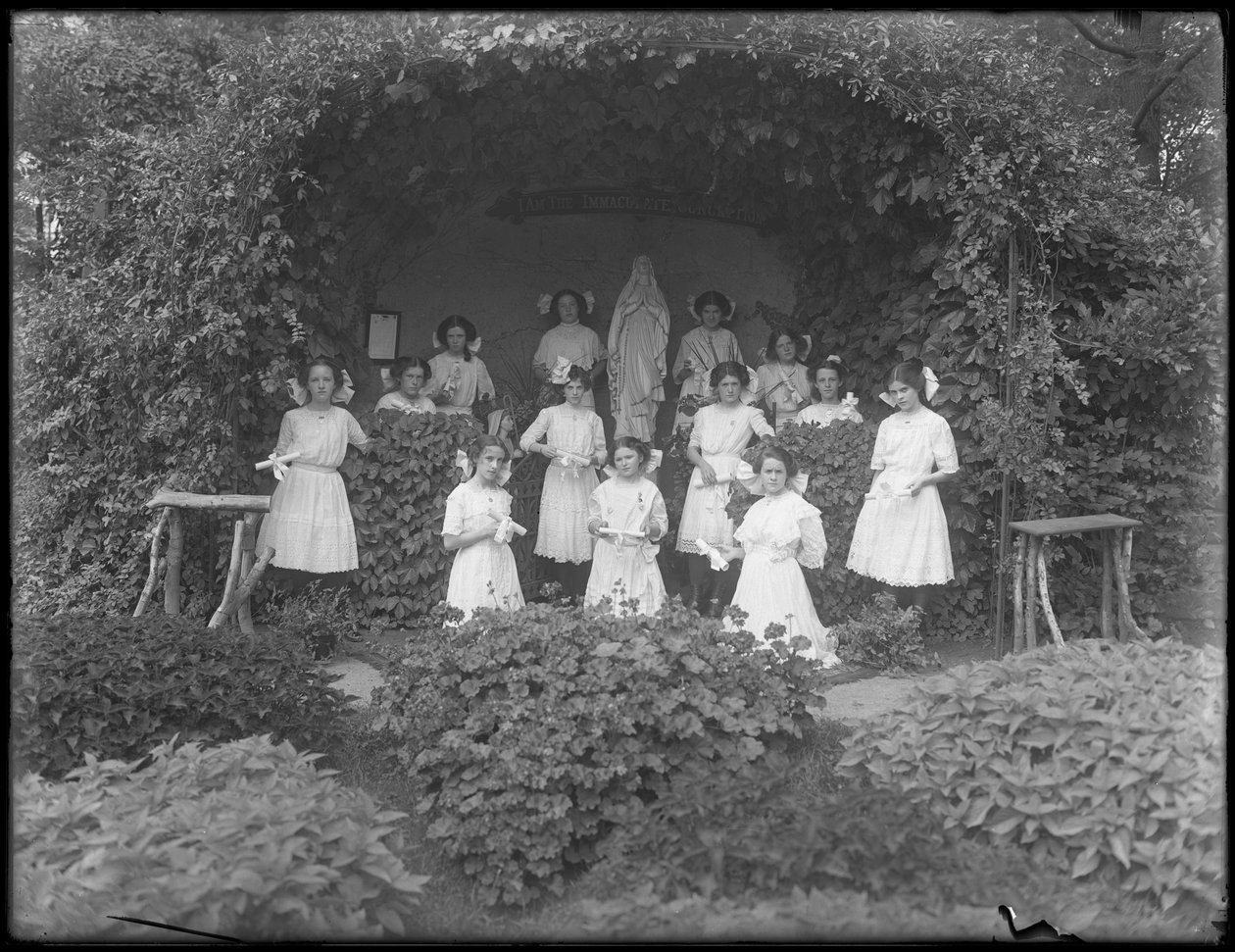 Afstudeerklas van meisjes van het Rooms-Katholieke Weeshuis poseert voor de grot met hun diploma