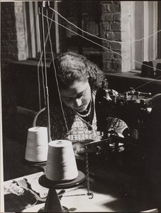 [Vrouw gebruikt een naaimachine met twee spoelen wit garen].