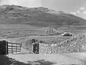 Zicht op de weg die over Hard Knott Pass gaat, twee mensen die bij een verkeersbord staan en een geparkeerde auto in de grasberm