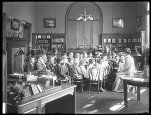 Kinderen luisteren naar een vrouw in de leeszaal van een niet-geïdentificeerde vestiging van de Queens Borough Openbare Bibliotheek, ca. 1910