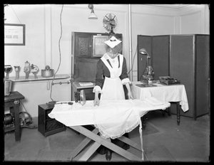 Jonge vrouw in een dienstmeisjeskostuum demonstreert het gebruik van een elektrische strijkijzer, ca. 1913-14