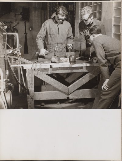 [Drie mannen met veiligheidsbrillen lassen een stuk metaal op een tafel]. door Arnold Eagle