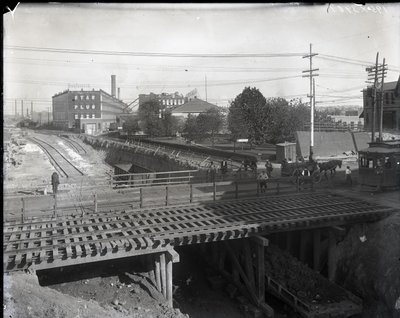 Horizontale zwart-witfoto van een houten brug en tramsporen over gedeeltelijk aangelegde spoorlijnen. Een tram gemarkeerd Chouteau 2137 begint de brug over te steken aan de rechterkant. In de verte fabrieksgebouwen met de tekst Star door Charles Clement Holt