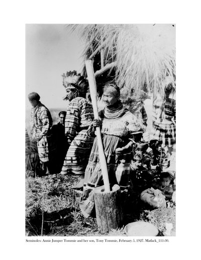 Annie Jumper Tommie gebruikt een vijzel en stamper om maïsmeel te maken, en haar zoon, Tony Tommie, draagt een adelaarsveren tooi, 5 februari, 1927 door Claude C. Matlack