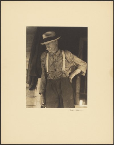 [Besnorde oude man met strohoed en bretels staat in de deuropening]. door Doris Ulmann