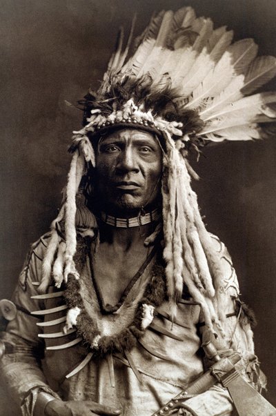 Weasel Tail (Apohsuyis). Portret van Indiaanse hoofdman van de Piegan stam met de beroemde adelaarsveer en wezels tooi, een suède tuniek, twee kettingen, een van bot en de andere van grizzly klauwen, en een tomahawk pijp door Edward Sheriff Curtis