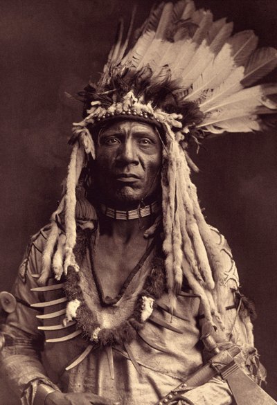 Weasel Tail (Apohsuyis). Deze Piegan Indiaan draagt de beroemde oorlogshoofdtooi van arendsveren en wezelskin, een suède tuniek, twee kettingen, een van bot en de andere van grizzlyklauwen, en een tomahawk pijp door Edward Sheriff Curtis