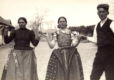 Twee dames en een man in Hongaarse traditionele kleding houden elkaars handen vast in een dans, met kaarsen in hun handen door Rudolf Balogh