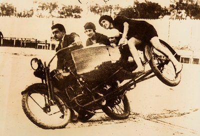 Een man pronkt met het rijden op een motorfiets met zijspan waarin drie jonge vrouwen zitten. Het zijspan komt van de grond bij het nemen van een bocht. door Unknown photographer