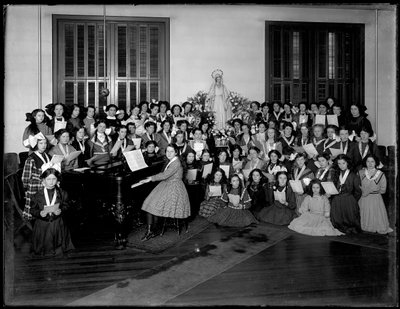 Groepsportret van een meisjeskoorles verzameld rond een piano met bladmuziek, waarschijnlijk in het Rooms-Katholieke Weeshuis in Kingsbridge, Bronx, ca. 1911 door William Davis Hassler