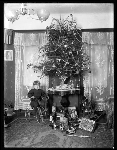 Seymour jongen poseert zittend op driewieler naast kerstboom in salon, Kerstmis 1912 door William Davis Hassler