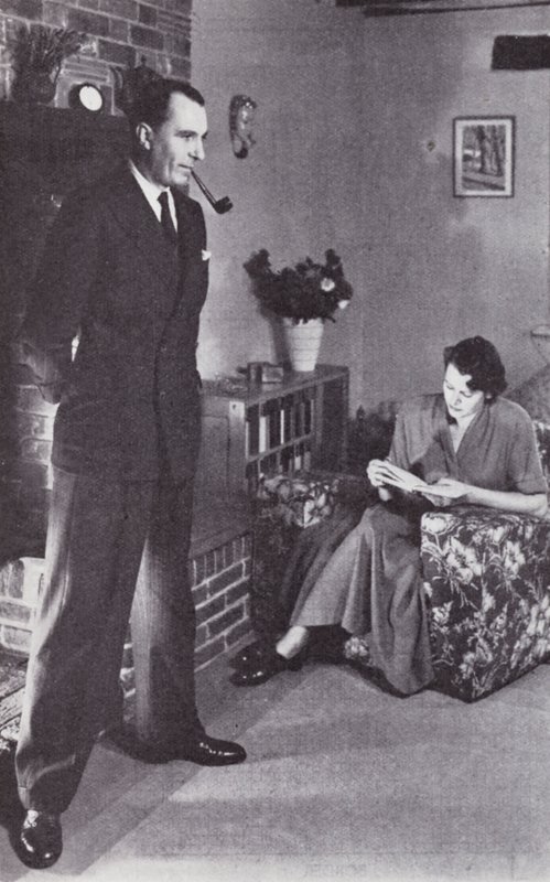 Vrouw leest, terwijl man zijn rug tegen het vuur verwarmt, ca. 1950 door English Photographer