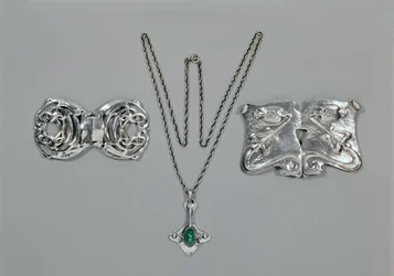 Zilveren sieraden ontworpen voor Liberty. Van links naar rechts: Taille gesp met verstrengeld Keltisch ontwerp, 1904. Hanger en ketting met cabochon geslepen chrysopraas. Taille gesp met repoussé ontwerp op gehamerd