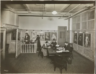 Hoofdkantoor/receptiekamer bij de Mutual Film Co. met secretaresse aan het bureau, mensen aan een tafel lezend, en portretten aan de muur, New York