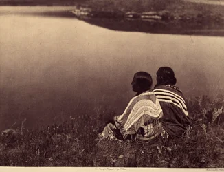 Een moment van rust: Piegan Indianen zittend aan de rand van een waterlichaam. Foto uit volume 6 van de encyclopedie gepubliceerd door Edward S. Curtis over de Indianen van Noord-Amerika, met een voorwoord van Theodore Roosevelt.