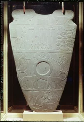 Het Narmer-palet: ceremonieel palet met koning Narmer, die de rode kroon van Beneden-Egypte draagt en geboeide en onthoofde gevangenen inspecteert, alsook een paar reusachtige langnekkige katten, gevonden in Hierakonpolis