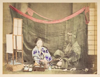 Japans stel in hun vrije tijd, Studiotableau van een jonge Japanse vrouw, zittend met theebenodigdheden om haar heen, die een pijp aanreikt aan een mannelijke figuur die achter haar zit, afgeschermd door een linnen muskietennet, Fotograaf: Onbekend Lucas C