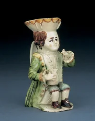 Toby-kan van een drinkebroer, zijn hoed met bruin-geel geschulpte rand, zijn ingekerfde bruine haar in een staart gebonden en met een witte rozet, zittend met een balustervormige kan op zijn knie, ca. 1770