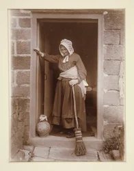 Een oude dame met een gistfles bij een cottage deur
