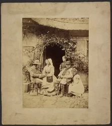 [Groep zittend voor open deur: twee mannen met hoeden, vrouw met muts, twee jonge meisjes - één staand].