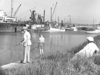 Een man en jongen vissen in Glasson Dock terwijl een achter hen zittende dame toekijkt, verschillende boten liggen afgemeerd