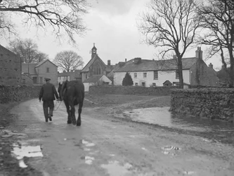 Man leidt paard over weg weg van camera naar dorpsgebouwen en kerk; architectuur; religieus; kerkelijk;