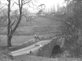 Twee meisjes zittend op een stenen brug over de rivier de Cocker aan de voet van Scale Hill, kijkend naar ganzen in de laan bij Loweswater