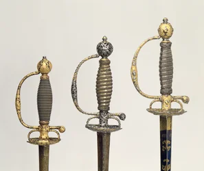 Heften van drie Franse zwaarden: een van staal, geblauwd en versierd met ontwerpen in gegraveerd goud, c.1730; een van staal, doorboord en gegraveerd en de grond van gematteerd goud, c.1760; een van gegraveerd staal