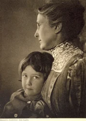 Halflang portret van een kind teder omhelsd door een oudere dame. De vrouw poseert met haar gezicht in profiel, terwijl ze de borst van de kleine jongen vasthoudt. Hij heeft een levendige en brutale blik, gericht op de camera