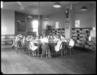 Kinderen luisteren naar een vrouw in de leeszaal van een niet-geïdentificeerde vestiging van de Queens Borough Public Library, ca. 1910