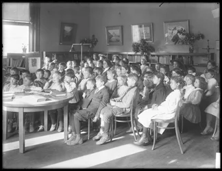 Kinderen zittend, luisterend in de leeszaal van een niet-geïdentificeerde vestiging van de Queens Borough Public Library, ca. 1910