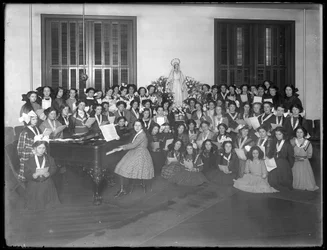 Groepsportret van een meisjeskoorklas verzameld rond een piano met bladmuziek, waarschijnlijk in het Rooms-Katholieke Weeshuis in Kingsbridge, Bronx, ca. 1911