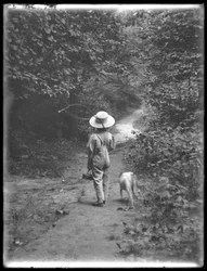 William Gray Hassler in strohoed en overall, met een visstok en mand, wandelend op een pad in het bos met een kleine hond (Bessie), 20 augustus 1911